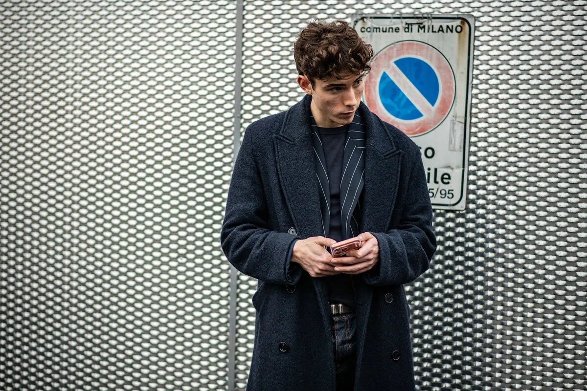 Реклама ChatGPT. Чоловік в елегантному темному пальто та піджаку в тонку смужку стоїть біля металевої огорожі з сітки, пильно дивлячись на екран свого телефону. Його зосередженість ідеально передає момент, коли користувач стикається з рекламою ChatGPT, адаптованою до його особистих уподобань. Це зображення символізує перетин класичної елегантності із сучасною алгоритмічною реальністю цифрового маркетингу.