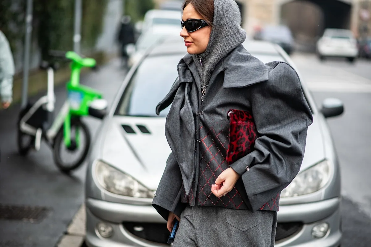 Шопінг у неділю, 25 січня 2026 року. На фото стильно одягнена жінка в сірому багатошаровому вбранні з курткою та сонцезахисними окулярами йде міською вулицею, на задньому плані — припаркований автомобіль та зелений скутер.