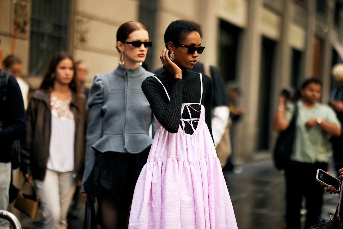 Стильні учасниці вуличного стилю на Тижні моди в Мілані – жінка в чорній водолазці та світло-рожевій сукні на бретельках, а також модель у сірому блейзері та чорних сонцезахисних окулярах, зняті в міській обстановці.