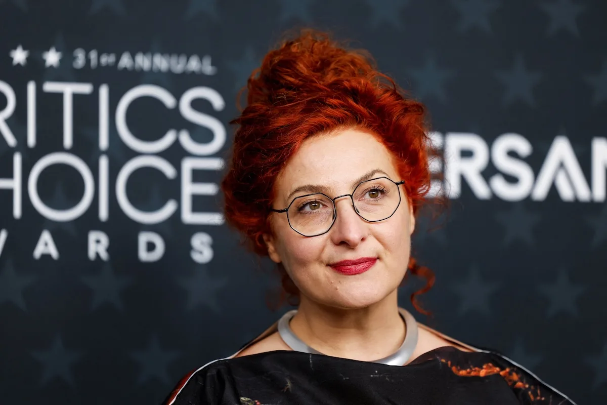 Польський художник по костюмах, номінований на «Оскар» на галузевому заході, портрет перед стіною церемонії вручення премії Critics’ Choice Awards, руде волосся та окуляри, елегантний стиль.