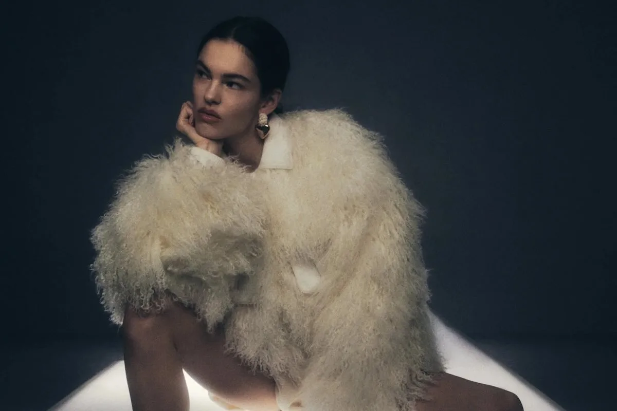 Модель у легкій шубі з архітектурним дизайном сидить у мінімалістичній студії, демонструючи сучасну жіночу естетику пошиття одягу преміального бренду.