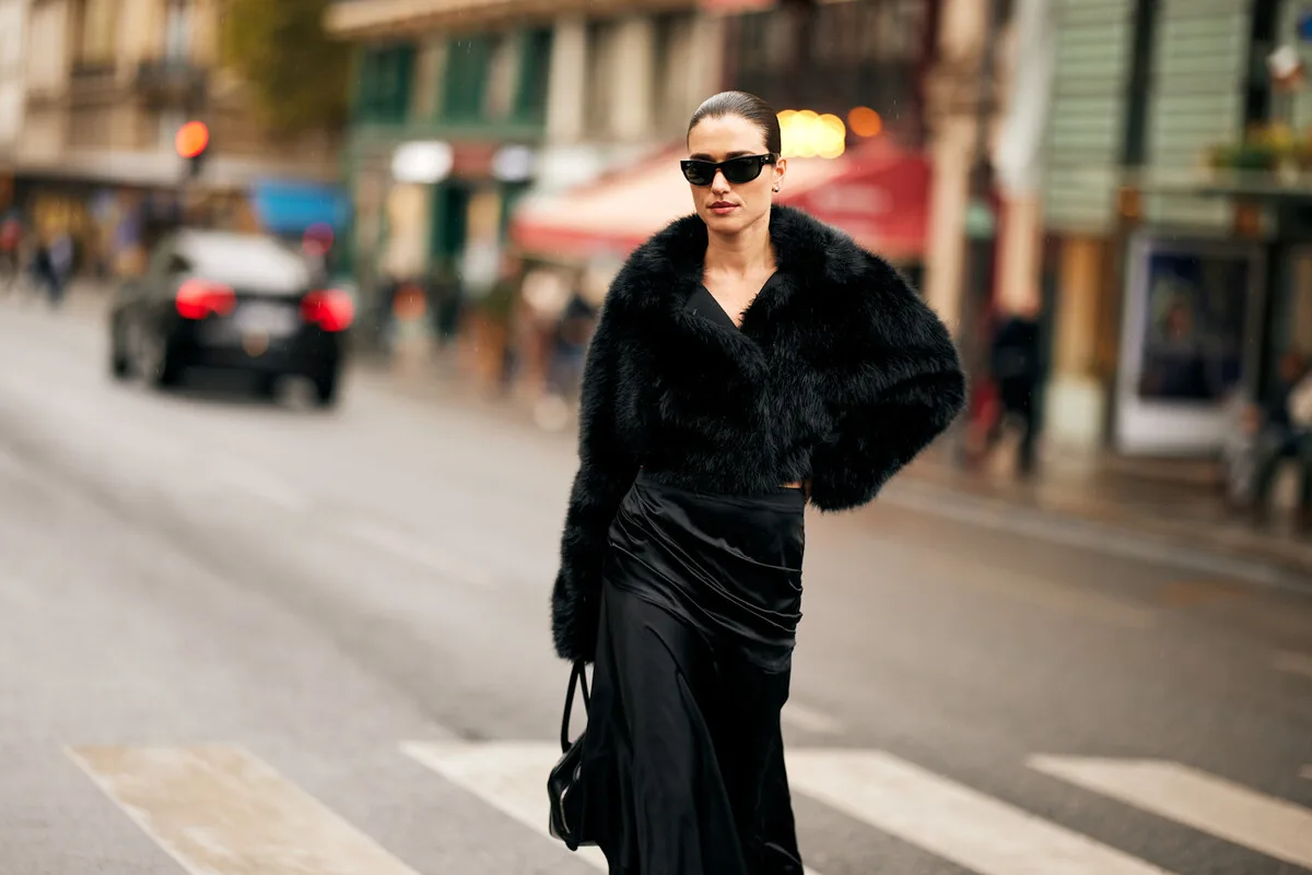 Saks Global Long. Жінка в елегантному чорному вбранні йде пішохідним переходом на міській вулиці. На ній коротка пухнаста шуба, атласна спідниця та темні сонцезахисні окуляри, а волосся гладко зачесане. На задньому плані видно розмитий рух транспорту, будівлі та вітрини магазинів, що надає роботі атмосфери столичного міста.