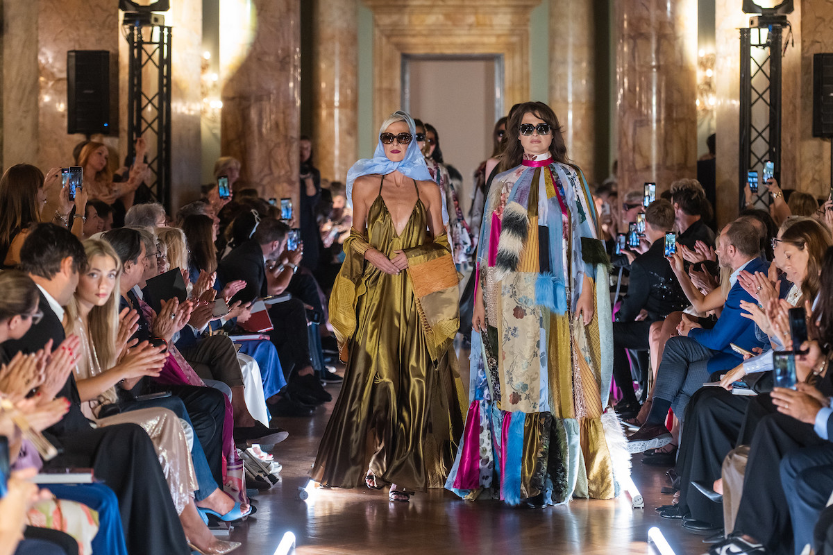 Дві моделі в довгих сукнях від кутюр, одна в золотому, інша в накидці з печворку, йдуть подіумом у розкішному залі.