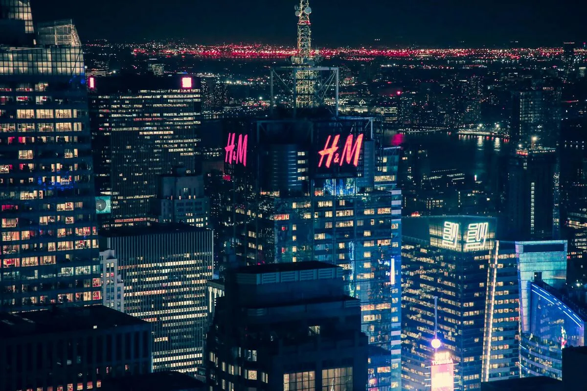 Фінансові результати H&M за 2025 рік. Хмарочос з логотипом H&M вночі, в центрі сучасного мегаполісу з підсвіченими офісними будівлями та міським горизонтом на задньому плані.
