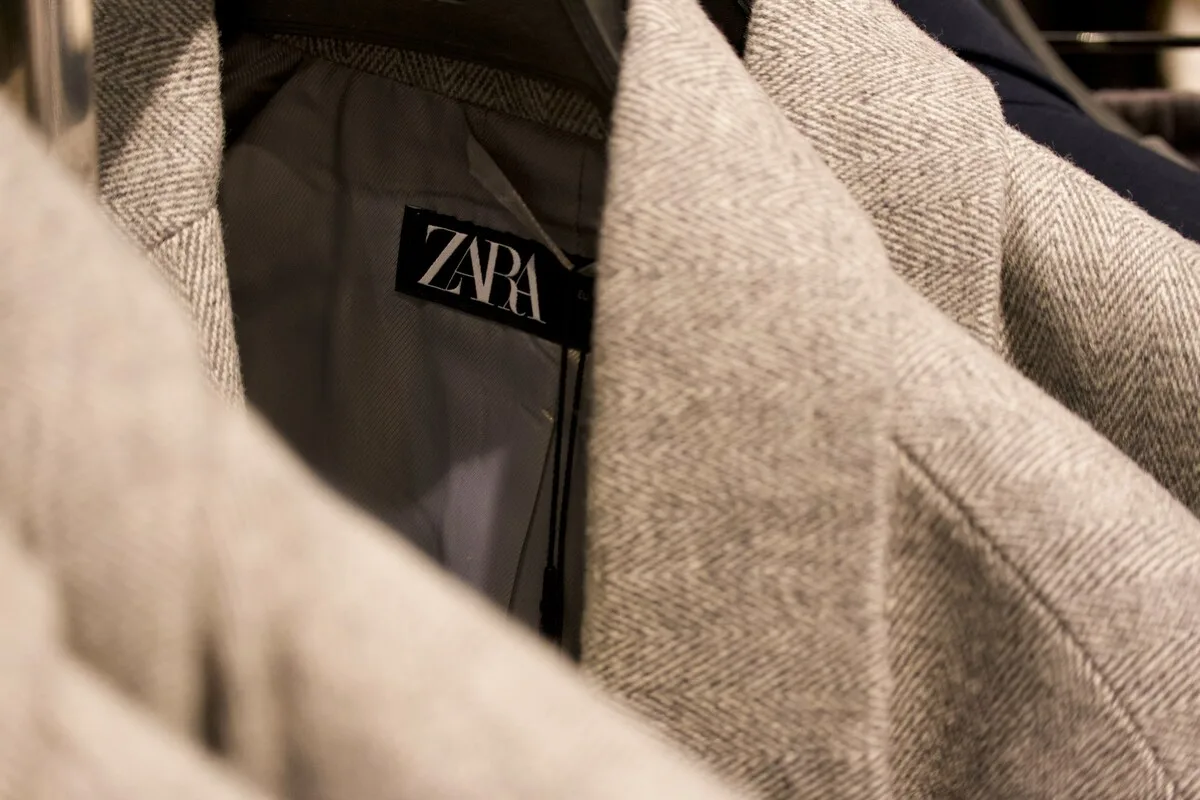«Зелений» маркетинг Zara від Управління з питань конкуренції та захисту прав споживачів. Деталь одягу в магазині: чорна етикетка ZARA, помітна на підкладці сірого пальто або куртки. Верхня тканина має чітке плетіння «ялинка». На задньому плані видно інший одяг на вішалках.