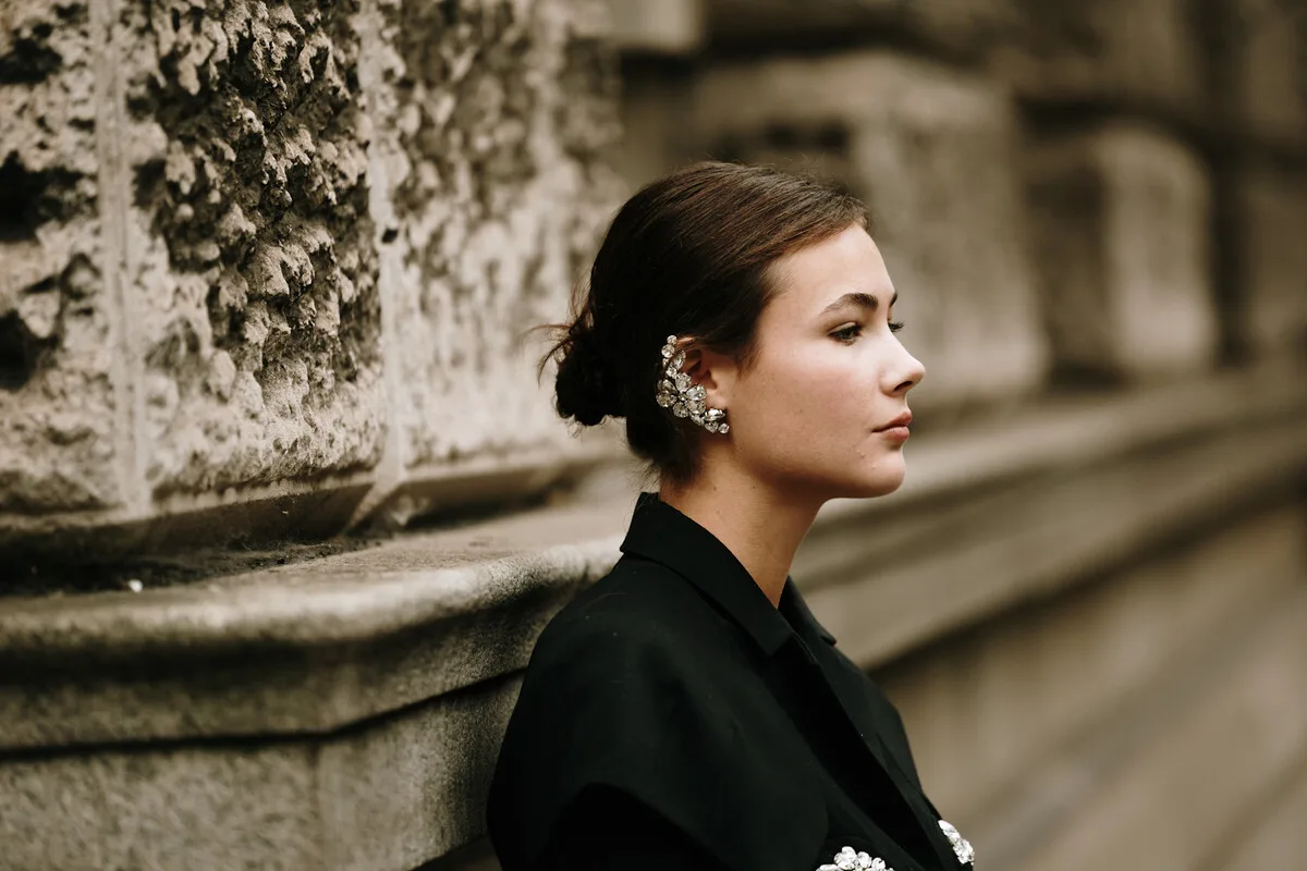 Ціна на срібло 2026. Портрет молодої жінки в профіль, що спирається на грубу кам'яну стіну; її волосся зібране в низький пучок, на ній чорна куртка та вишукана блискуча срібна сережка, а фон вулиці розмитий.