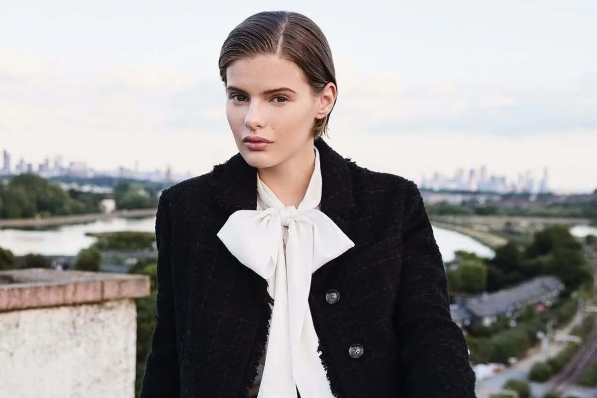 Л. К. Беннетт зачиняє магазин. Молода жінка в чорному пальто та білій блузці, зав'язаній бантом, стоїть на тлі річки та міського пейзажу, дивлячись прямо в камеру.