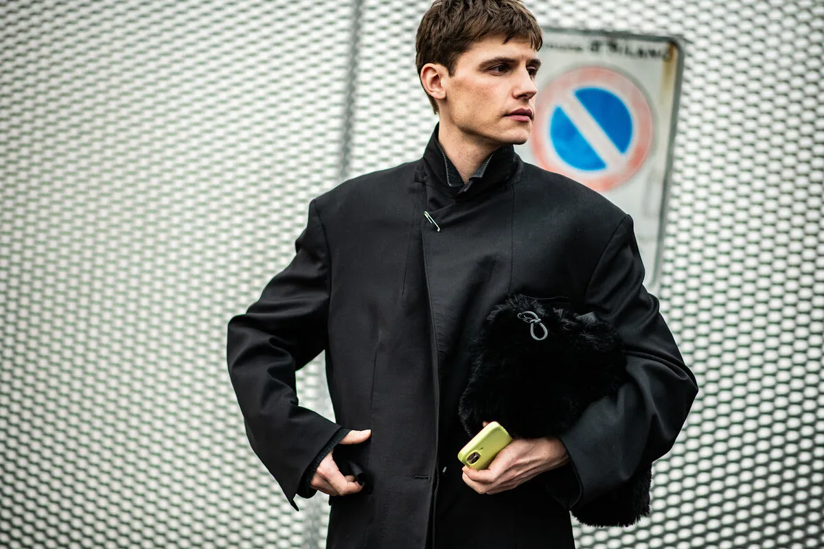 Грінвошинг від Tchibo, Bolt. Портрет чоловіка в мінімалістичному чорному пальто зі стійкою, застібнутим на шиї шпилькою. Він тримає пухнасту чорну сумку та жовтий телефон під пахвою, стоячи на тлі металевої сітки та дорожнього знака.