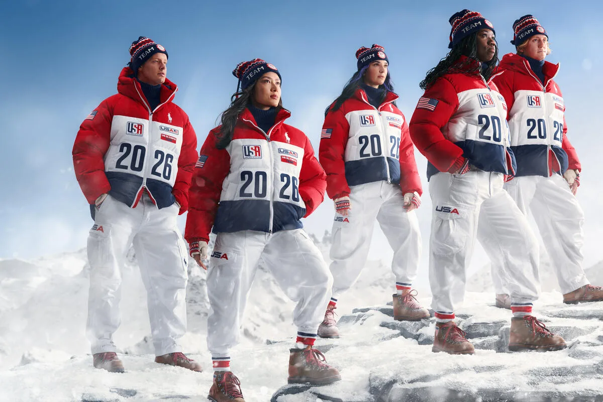 Розкішні бренди на Олімпійських іграх 2026 року в Мілано-Кортіна. Група спортсменів збірної США у своїй зимовій олімпійській формі, одягнених у червоно-біло-сині куртки та білі штани з написом 2026, стоять на засніженій гірській місцевості, дивлячись у далечінь.