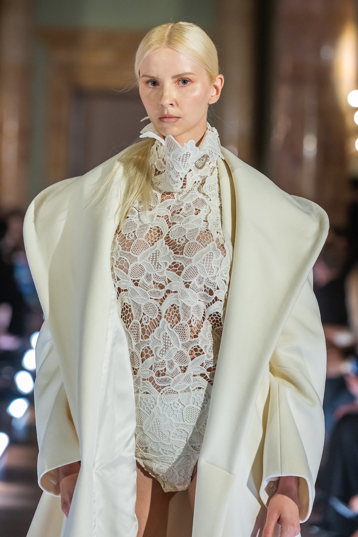 Модель у білому мереживному боді з високим коміром та кремовому пальто під час показу мод. Східний напрямок до Риму.