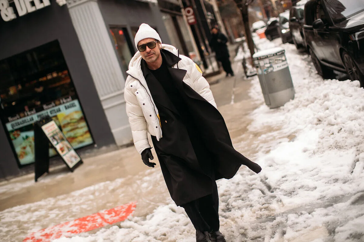 Куртки PFAS від C&A. Чоловік у білій пуховику, довгому чорному пальто, білому капелюсі та сонцезахисних окулярах йде засніженим тротуаром у міському середовищі. Вулиця зимова, на задньому плані припарковані машини, сльота та магазини. Фігура виглядає стильно та впевнено, коли йде до камери.