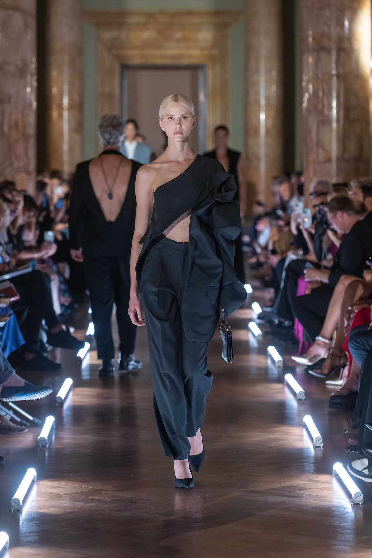 Дизайн Моніки Суровєц. East Bound Roma – польський показ мод у Римі: модель йде подіумом у чорній асиметричній сукні з відкритими плечима та елегантних штанах.