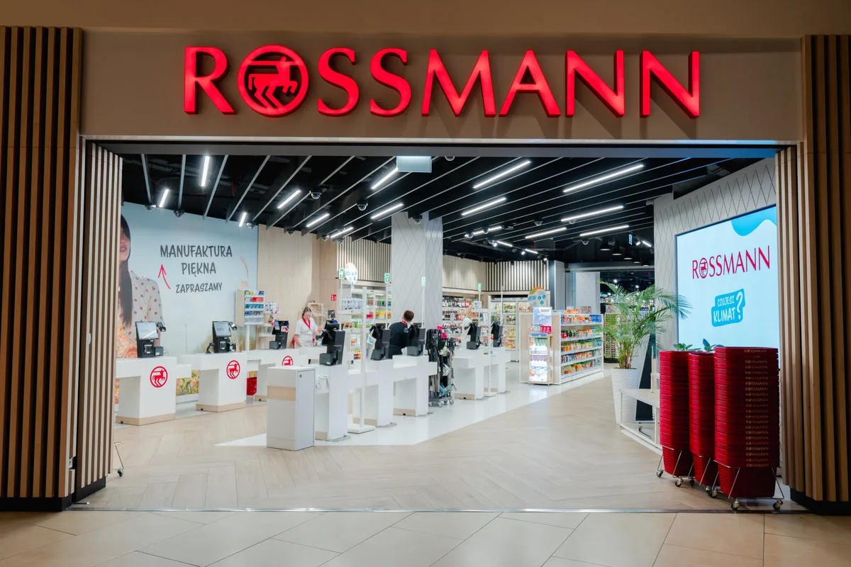 Інтернет-аптека Rossmann. Вид на вхід до сучасної аптеки з логотипом Rossmann, касами самообслуговування, полицями з продуктами та червоними кошиками для покупок.