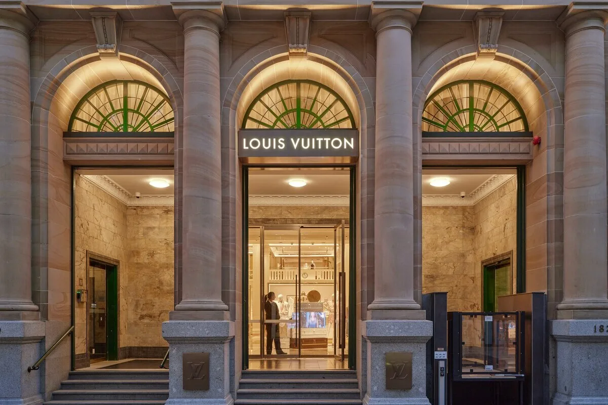 Louis Vuitton карає відмивання грошей. Вхід до бутіку Louis Vuitton, розташованого в елегантній кам'яній будівлі з високими колонами та арочними вікнами, прикрашений підсвіченою вивіскою бренду над скляними дверима, а всередині — яскравим, розкішним інтер'єром з вітриною сумок.