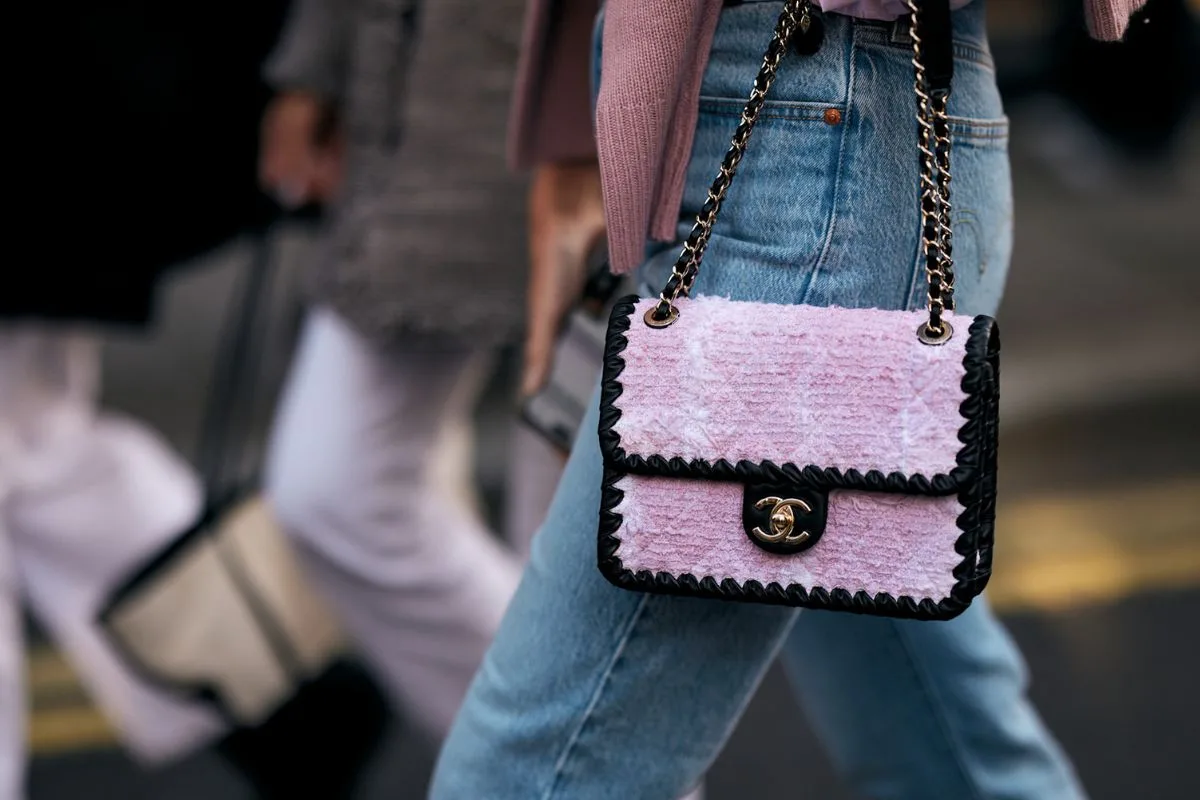 На фотографії зображено образ у вуличному стилі з Лондонського тижня моди, де жінка одягнена в сині джинси з високою талією та світло-рожевий светр. Центральним елементом композиції є розкішна сумка Chanel з текстурованого рожевого твіду з чорною окантовкою та золотою застібкою. Ця деталь підкреслює високу якість преміальних аксесуарів, на яку можуть вплинути нові податкові правила у фешн-індустрії. Фон трохи розмитий, що показує динамічний міський рух та інших стильних персонажів.