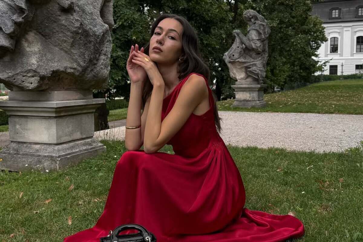 Український бренд Vovk залишає польський ринок. Жінка в довгій червоній сукні сидить на траві в оточенні кам'яних скульптур та зелені, тримаючи при собі елегантну чорну сумку.