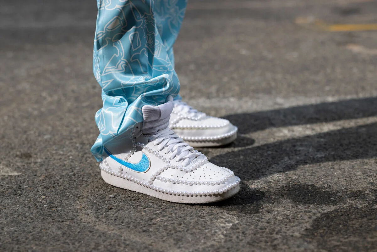 Культові бренди кросівок 2026 року за версією RetailBoss. Білі кросівки Nike зі світло-блакитним логотипом, прикрашеним перлами навколо підошви та верху, у поєднанні з синіми штанами з візерунком на асфальтовій поверхні.