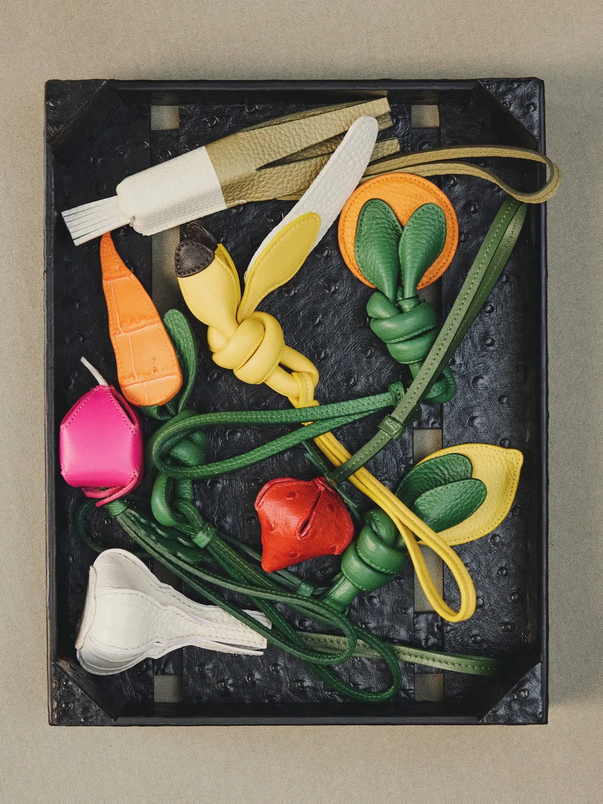 Шкіряні брелоки Jacquemus у формі моркви, банана, лимона та вишні, розташовані на чорному підносі.