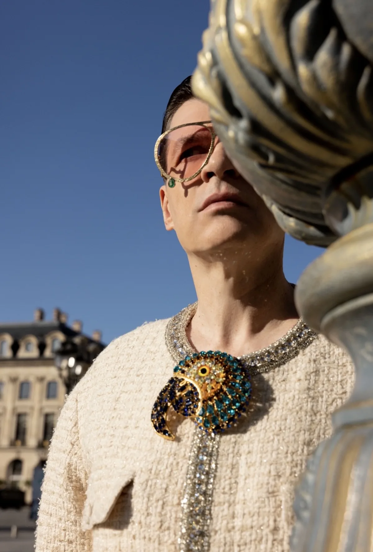 Крупний план чоловіка у світлому твідовому костюмі з великою, вишуканою брошкою у формі птаха та рожевими сонцезахисними окулярами, що стоїть біля декоративного ліхтарного стовпа на тлі класичної архітектури.