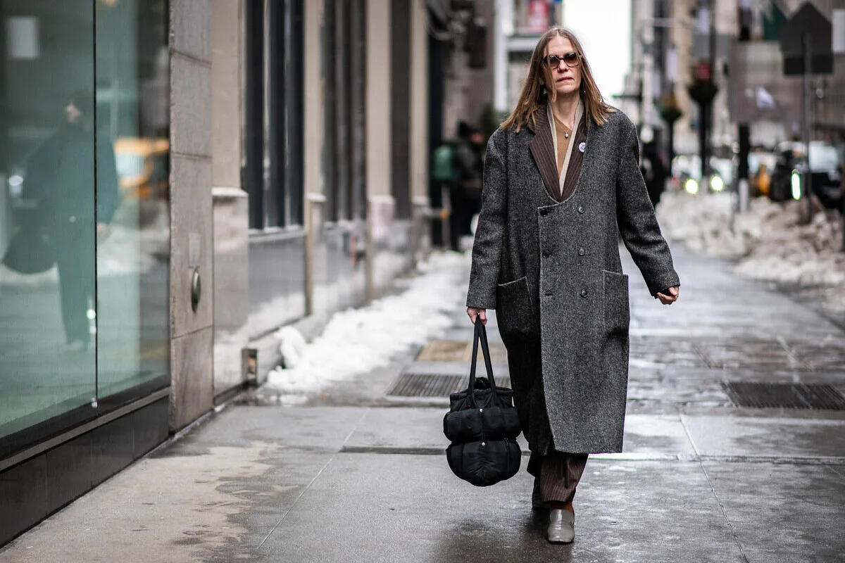 Чи 15 лютого 2026 року – це неділя шопінгу? Жінка йде вулицею в довгому вовняному пальто графітового кольору, яке поєднує з класичними штанами та мінімалістичним взуттям. Чорна стьобана сумка та сонцезахисні окуляри доповнюють образ. Цей міський образ з його стриманою кольоровою палітрою відповідає зимовій естетиці повсякденного вуличного одягу.