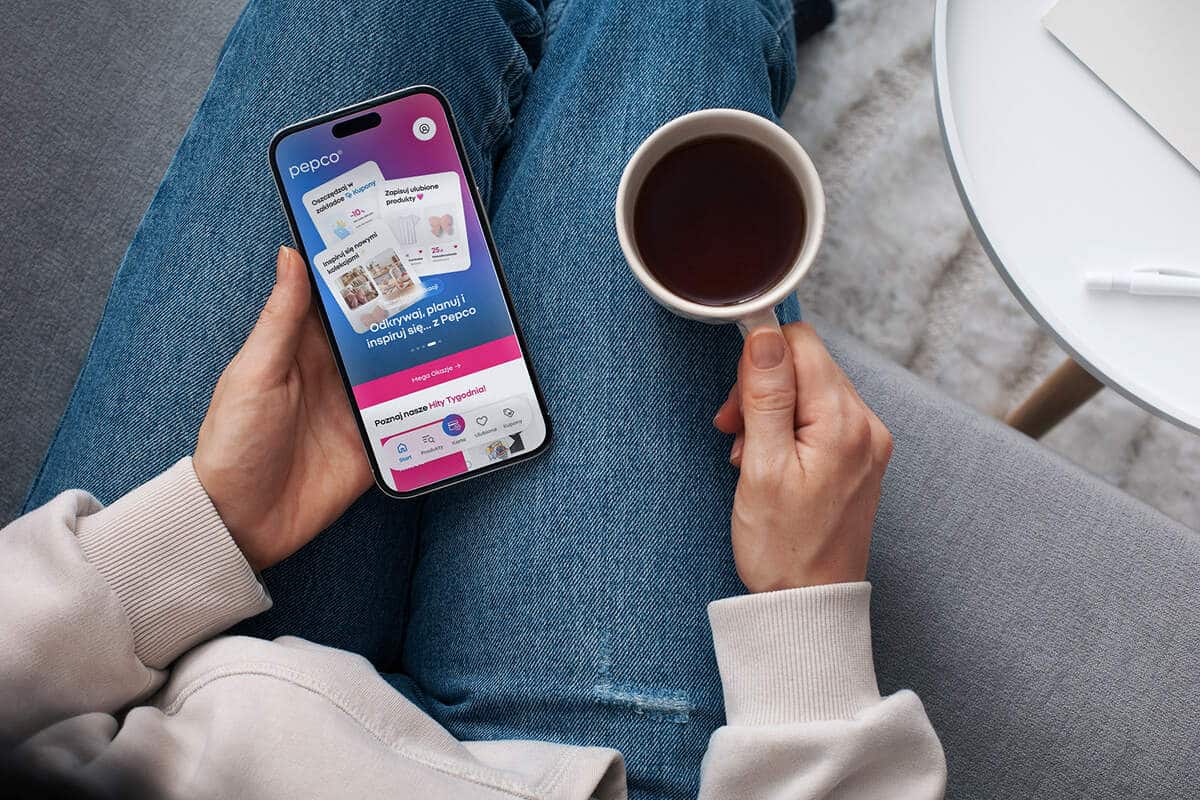 Мобільний додаток Pepco Polska. На фото: Візуалізація додатку Pepco на смартфоні в руках жінки, яка сидить на дивані з чашкою кави.