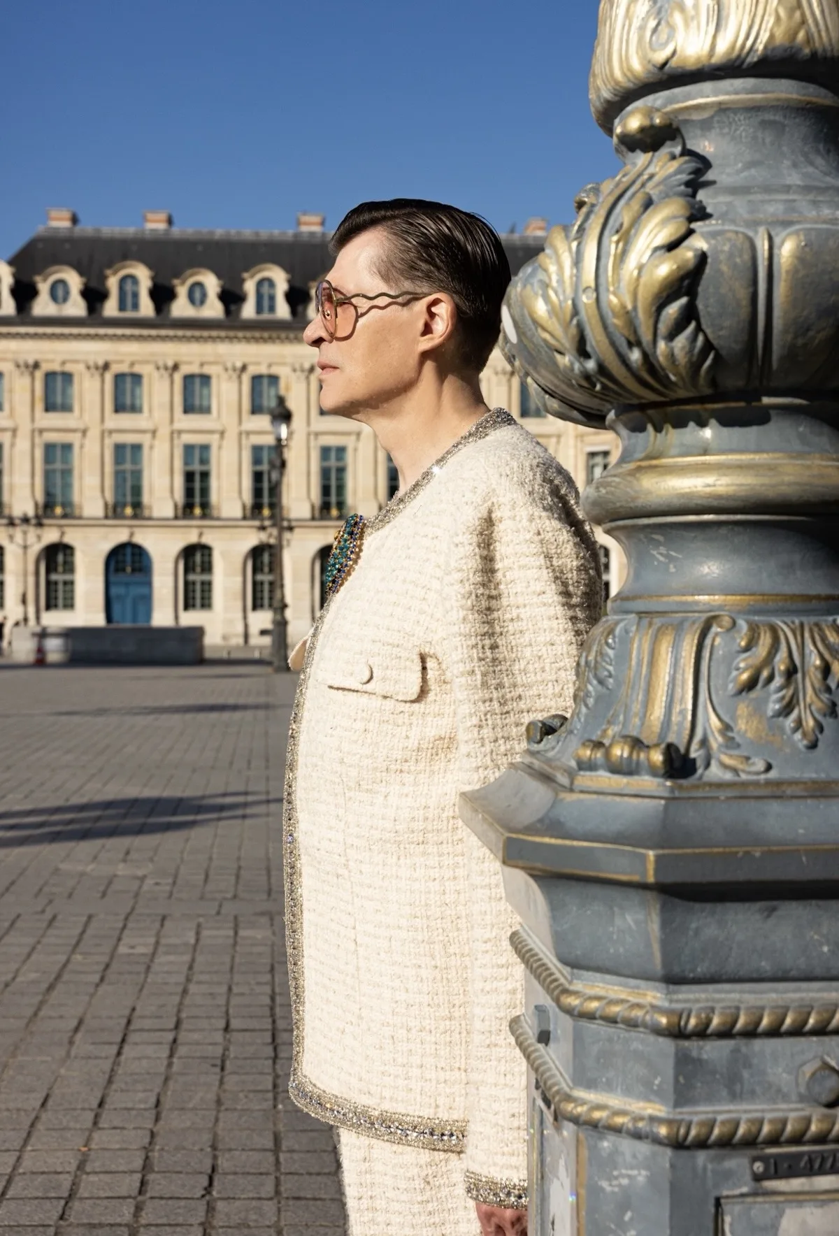 Профіль чоловіка в елегантному світлому костюмі з мерехтливою обробкою, який стоїть поруч із вишуканим ліхтарним стовпом на міській площі з історичною будівлею на задньому плані.