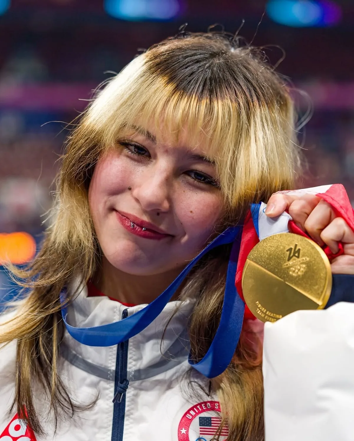 Стиль Аліси Лью помітний у деталях — природному макіяжі, неповторній зачісці та прикрасах, які підкреслюють її індивідуальний образ. На фотографії фігуристка демонструє свою золоту медаль, тримаючи її близько до обличчя та посміхаючись у камеру. Крупний план передає емоції перемоги та щирість, що стали візитною карткою її повернення.