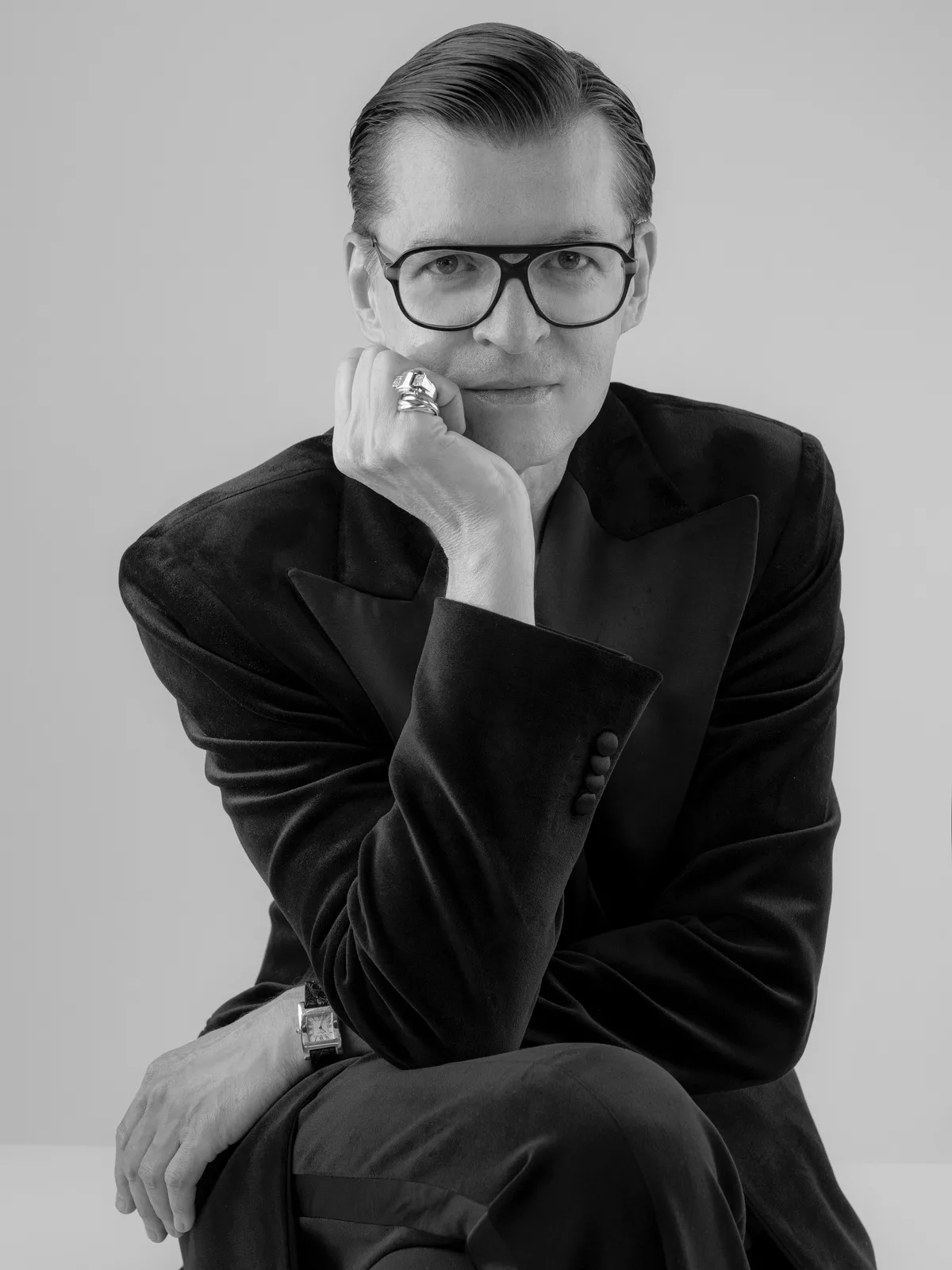 Адам Лея. Чорно-білий портрет чоловіка в окулярах, одягненого в елегантний темний піджак, який поклав голову на руку та дивиться прямо в камеру.