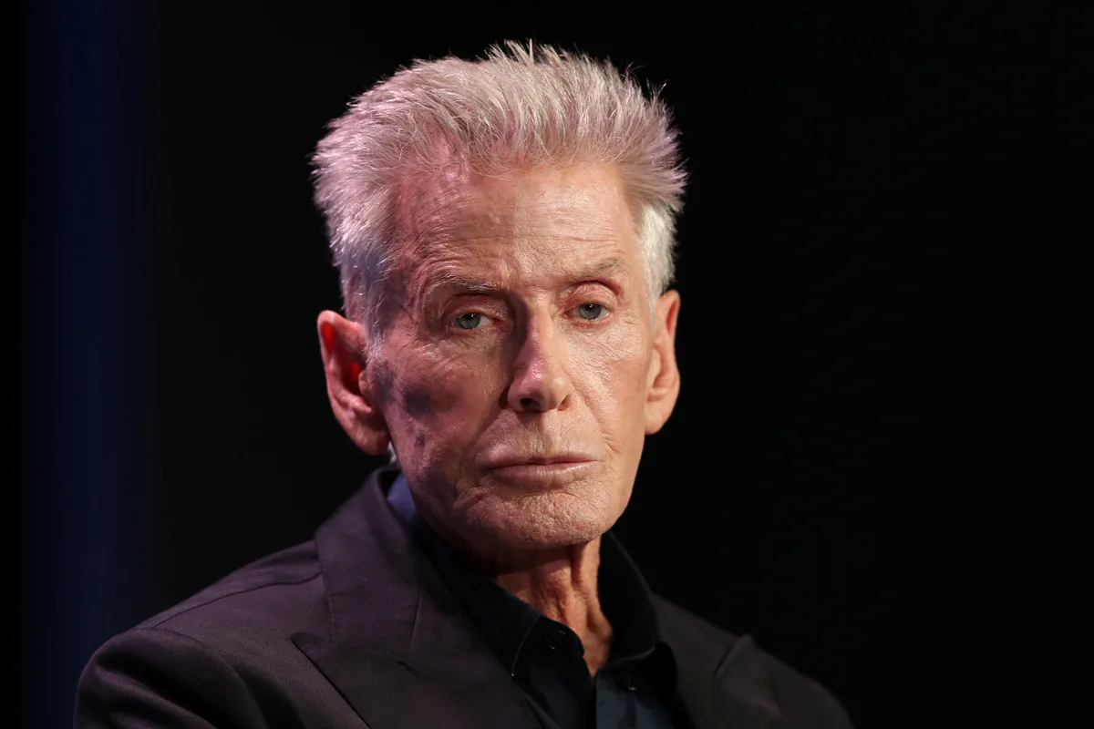 Дружина Кельвіна Кляйна. Літній чоловік з коротким сивим волоссям та чіткими рисами обличчя сидить на темному тлі, одягнений у чорний піджак та темну сорочку, освітлений прожектором, із серйозним виразом обличчя.