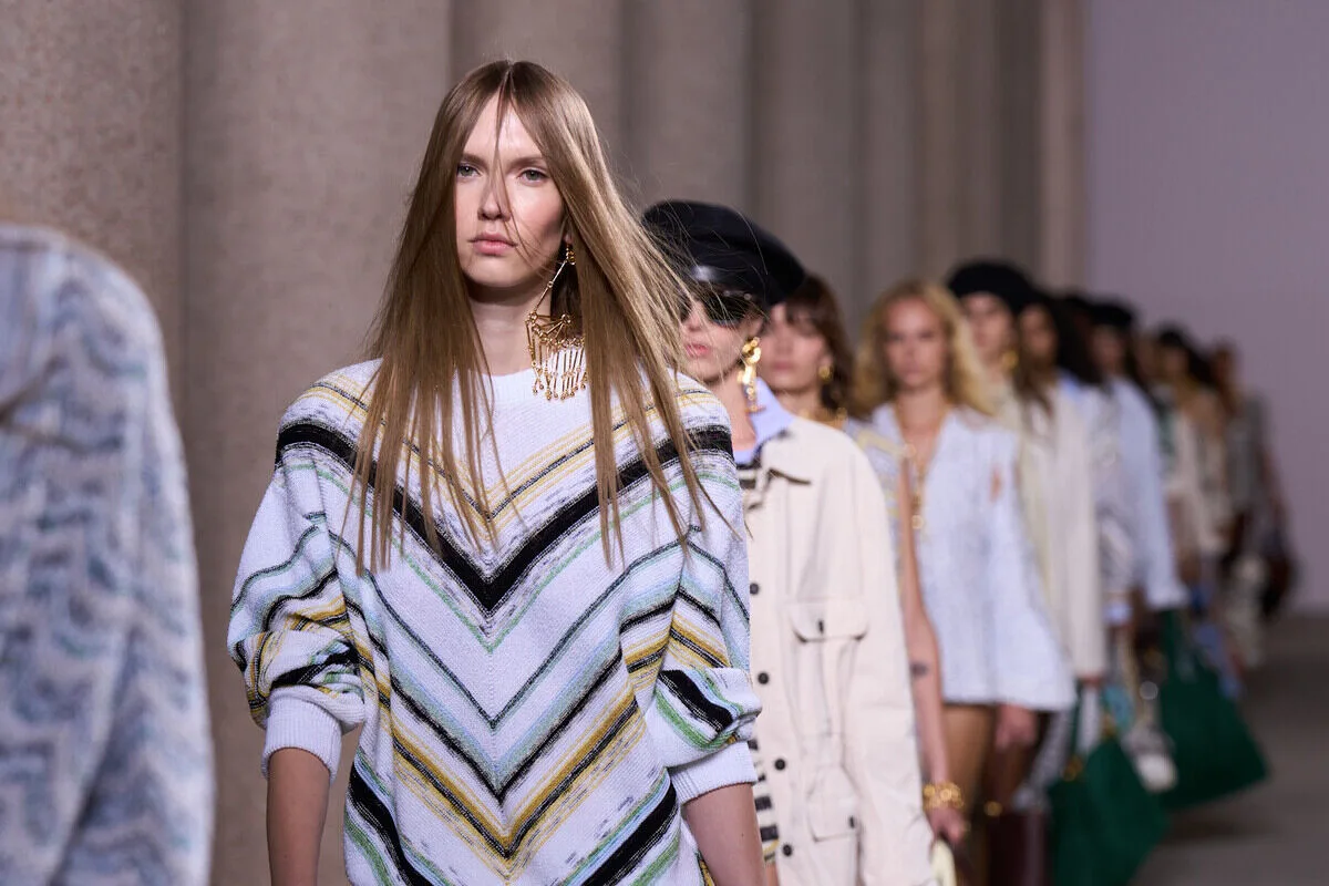 Захоплення Missoni. Модель йде подіумом у в'язаній сукні або светрі з характерними зигзагоподібними смужками яскравих кольорів, а інші моделі в схожих стилях йдуть позаду неї під час показу мод в елегантному просторі з колонами.