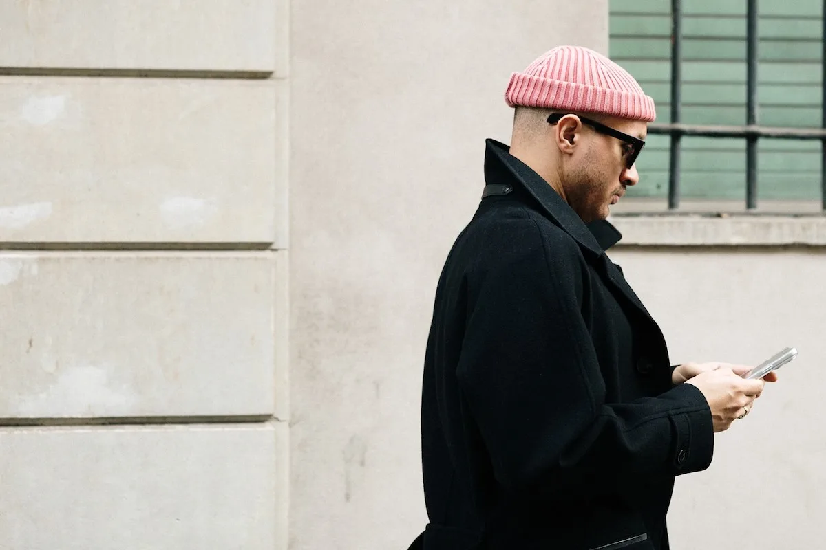 Чоловік у чорному пальто та рожевій шапці-біні користується смартфоном на вулиці – користувач соціальних мереж та онлайн-майданчика.