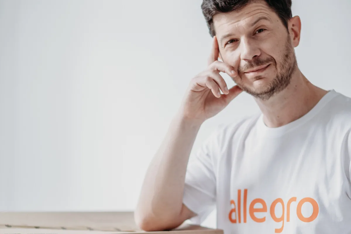 Послуги Allegro. На фото: Марцін Кусьмєж, генеральний директор Allegro, у футболці з логотипом компанії, спираючись на картонну коробку з вантажем.