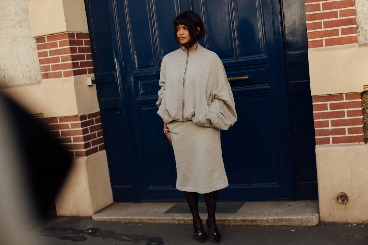 Модель у мінімалістичному сірому вбранні від H&M — оверсайз-толстовці та спідниці міді — стоїть перед темно-синіми дверима багатоквартирного будинку.