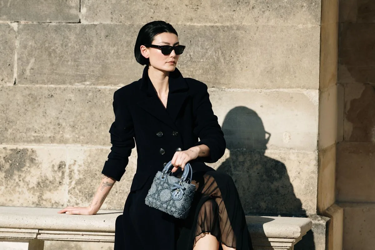 Жінка в елегантному чорному пальто та темних окулярах сидить на кам'яній лавці, тримаючи в руках розкішну сумочку. Цей образ ідеально підходить для офіційних подій та весняних прогулянок, поки ми розмірковуємо, чи є Страсна п'ятниця 2026 року вихідним днем і як стильно спланувати святковий сезон.