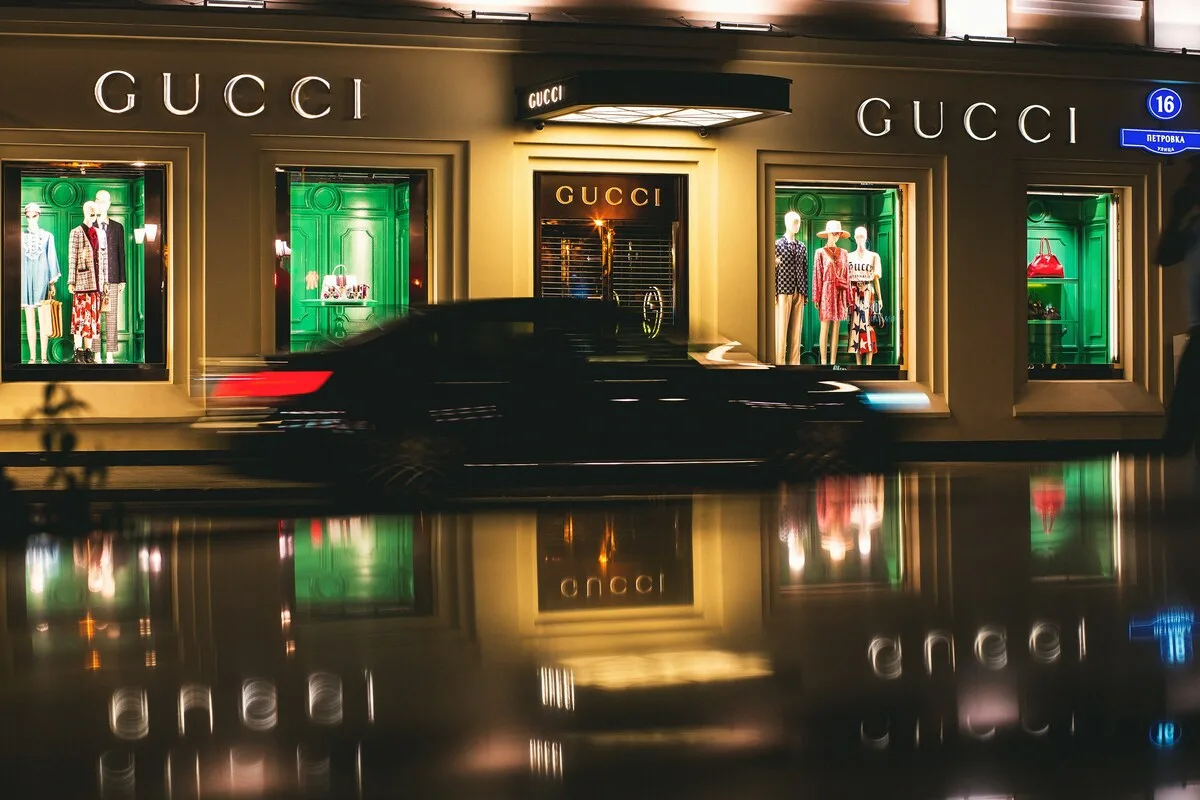 Розкіш на Близькому Сході 2026. Розкішний бутік Gucci вночі з підсвіченими вітринами та елегантною вітриною одягу та сумок відбивається на мокрій вулиці, а на передньому плані розмита картина автомобіля, що проїжджає повз, представляє ексклюзивний магазин модного одягу в центрі міста.