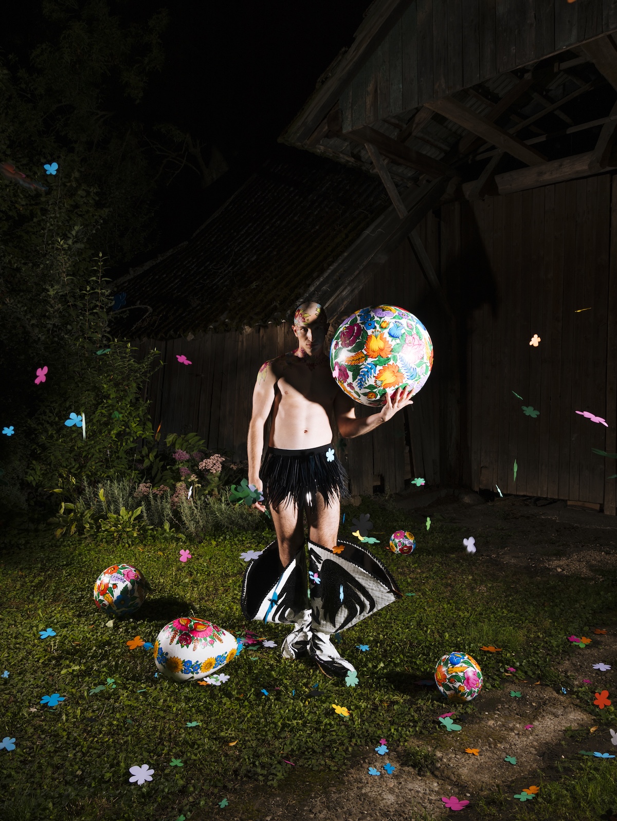 Модель стоїть перед дерев'яною будівлею вночі, тримаючи велику кулю з квітковим візерунком. Навколо розкидані декоративні розписні яйця та різнокольорові паперові квіти, а сама модель одягнена в чорну спідницю з бахромою та високі чоботи.