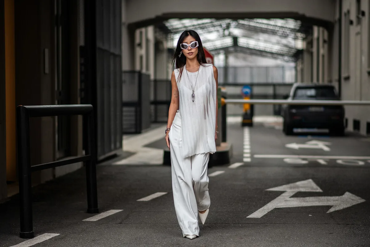 Прем'єра серіалу «Пластикова детоксикація» від Netflix, про що вона? Жінка йде вулицею в сучасному міському середовищі, одягнена в елегантний білий ансамбль, що складається з вільної блузки та штанів широкого крою. Вона в яскравих сонцезахисних окулярах і виглядає як модель на модній фотосесії. На задньому плані видно парковку та промислову архітектуру міста.