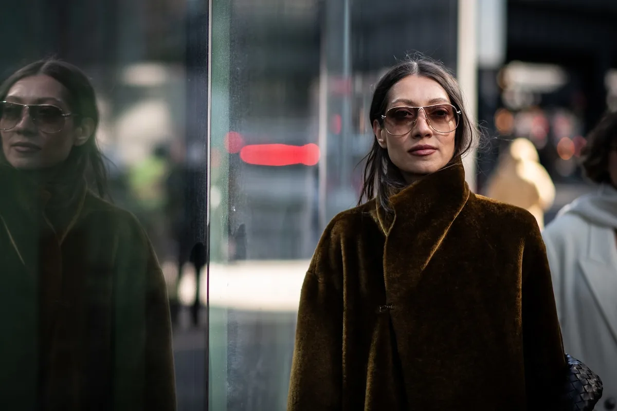 Жінка в мінімалістичному розкішному пальто стоїть біля скляної вітрини, демонструючи сучасний міський стиль. Купівля такого одягу на іноземних платформах може змінитися, оскільки Європейський Союз планує запровадити плату за обробку відправлень з-за меж ЄС.