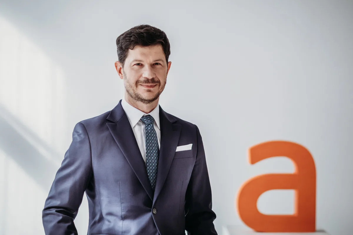 Фінансові результати Allegro за 2025 рік. Портрет генерального директора Allegro в елегантному темно-синьому костюмі та краватці, який стоїть у світлому, сучасному офісному приміщенні. Він злегка посміхається та дивиться в камеру, а на руці — велика помаранчева літера.