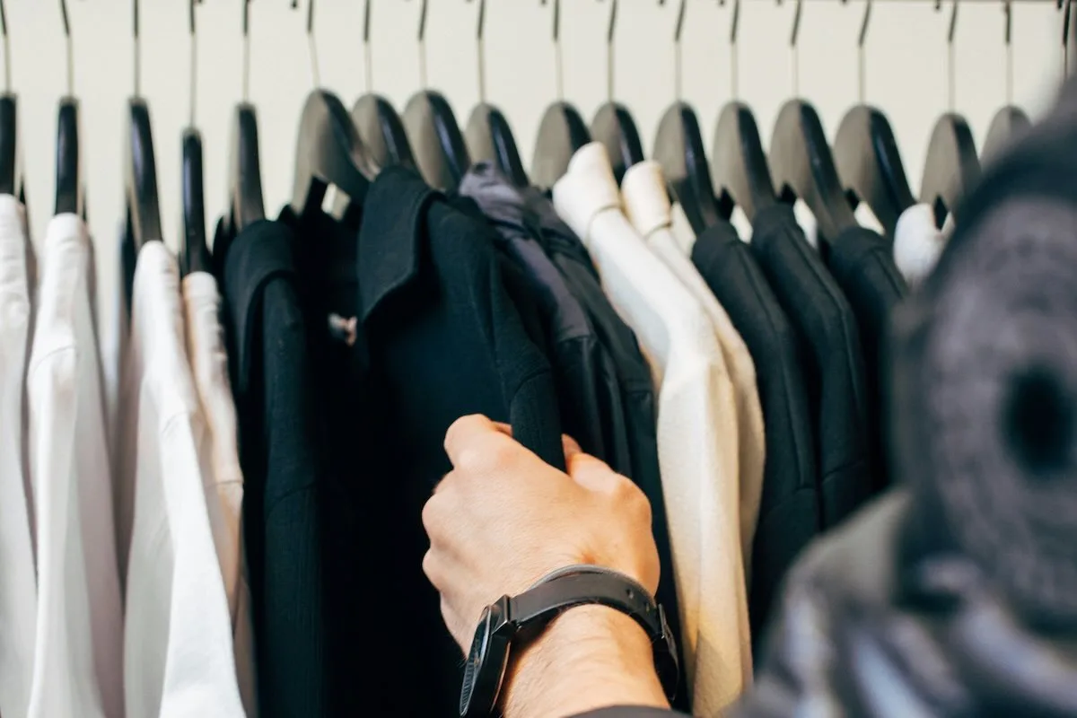 Чи відкриті торгові центри у Страсну п'ятницю 2026 року? На фотографії зображена людина, яка переглядає одяг на вішалках у шафі, вибираючи чорну сорочку серед інших речей приглушених кольорів.