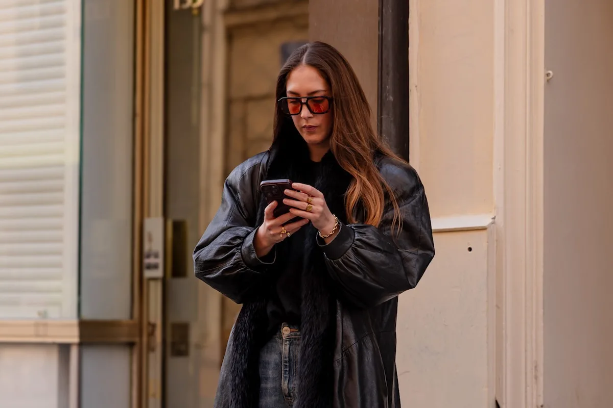 Технологічні тренди, які трансформують індустрію моди у 2026 році. Жінка в чорному шкіряному пальто та великих сонцезахисних окулярах стоїть на вулиці та користується смартфоном.