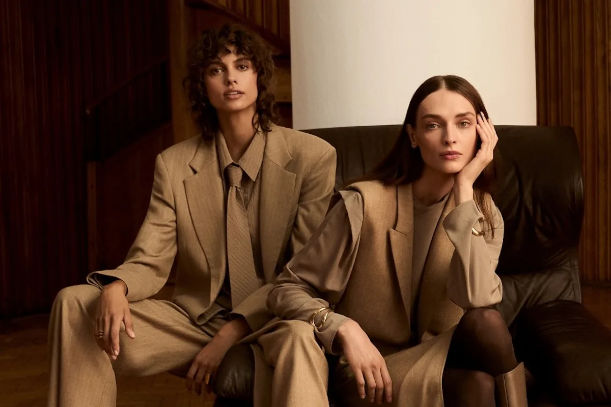 Дві моделі в бежевих костюмах JEMIOL сидять на шкіряному дивані в елегантному дерев'яному інтер'єрі.