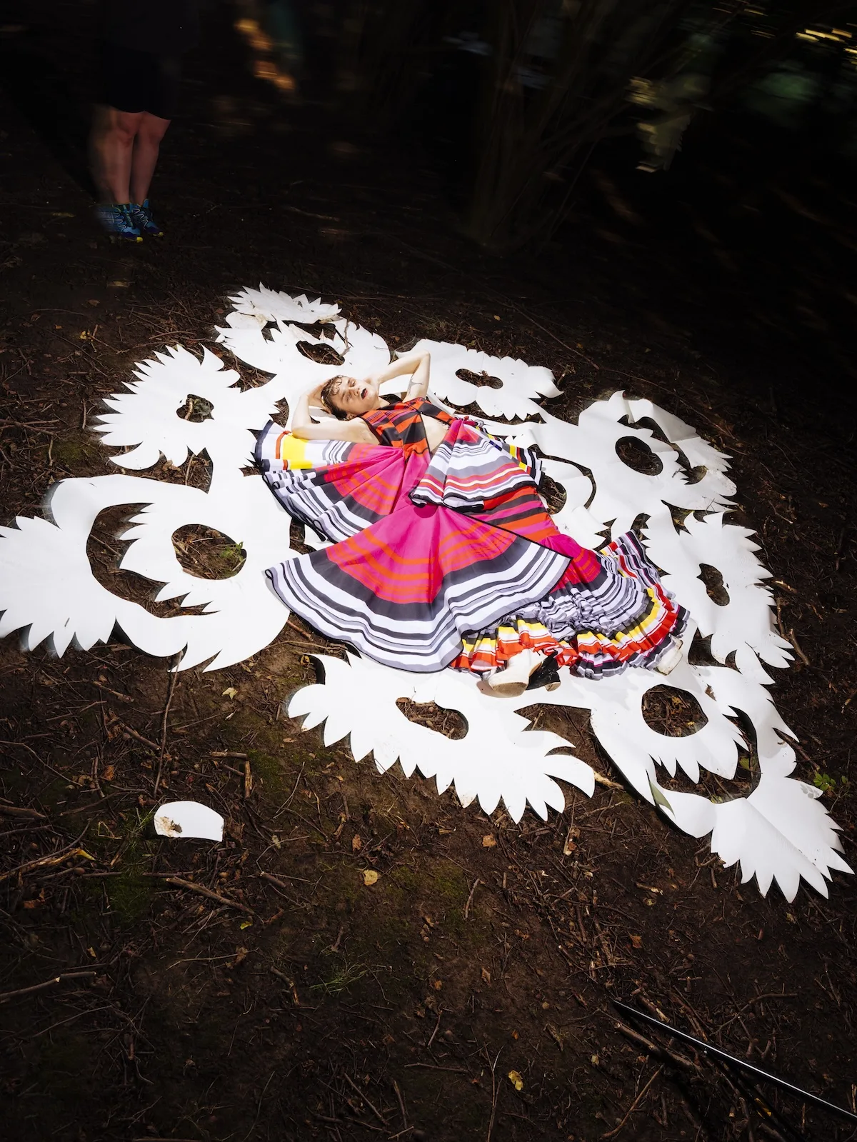 Модель лежить на землі в лісі на білій вирізаній інсталяції з квітковими та пташиними мотивами. На ній широка різнокольорова сукня з хвилястою спідницею та насиченими смужками.