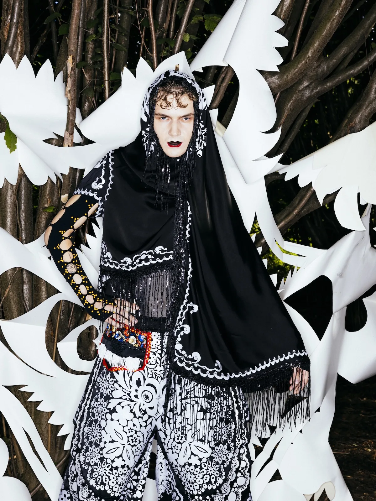 Модель стоїть серед дерев на тлі білих різьблених фігур, що нагадують птахів та квіти. На ній чорний вишитий одяг з бахромою та візерунками в фольклорному стилі, а також головний убір з чорною хусткою.