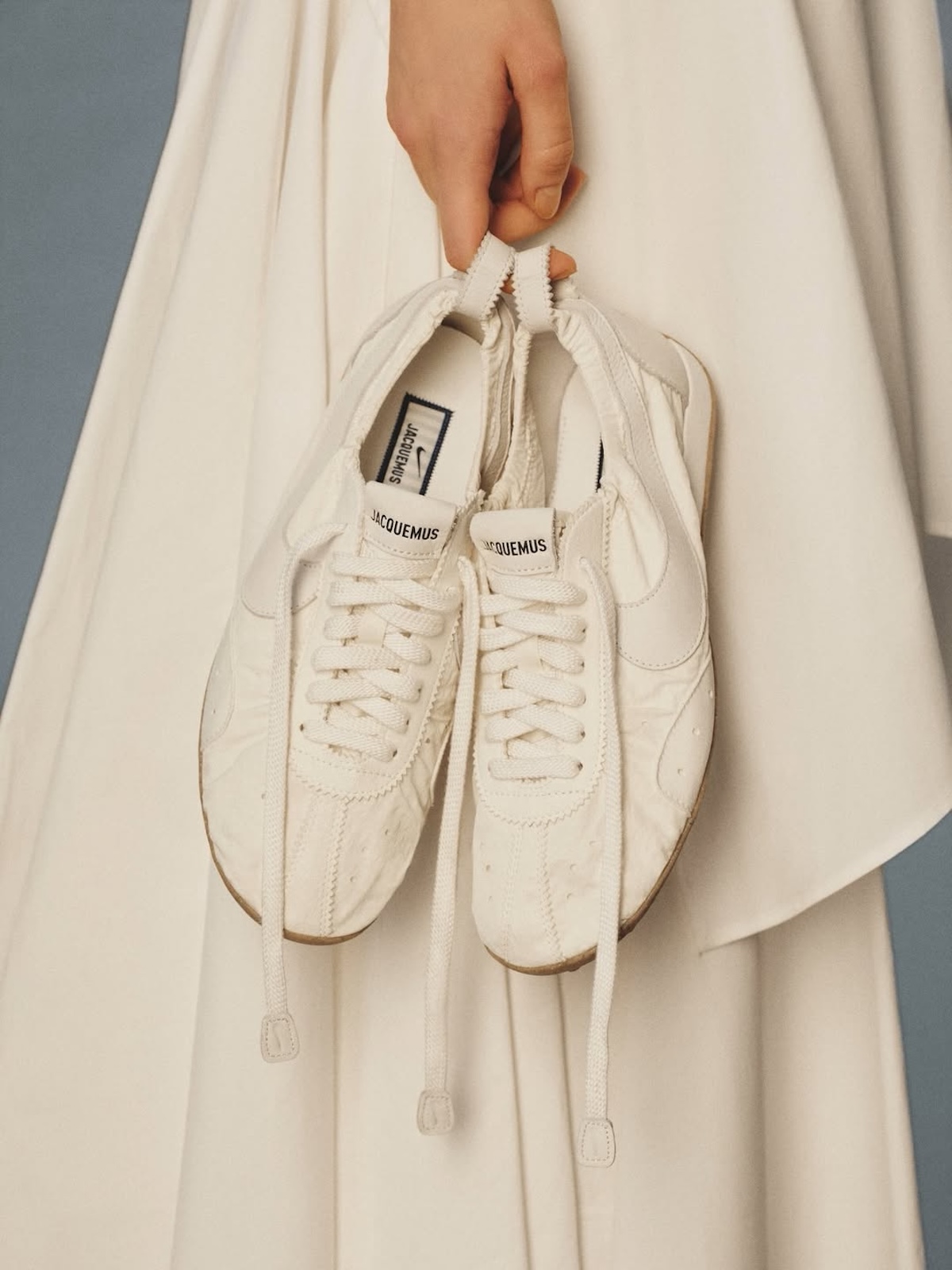 Кросівки Jacquemus x Nike, що тримаються в руці на тлі легкої кремової тканини, демонструючи їхню ніжну конструкцію та видовжені шнурівки. Колекції кросівок весни 2026 року все більше схиляються до елегантності та простоти, переосмислюючи класичні моделі в більш модному контексті.