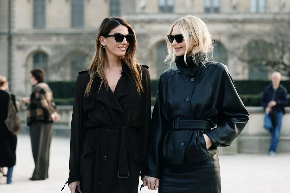 Дві елегантні жінки в темних сонцезахисних окулярах прогулюються на тлі класичної європейської архітектури. Одна одягнена в легкий чорний тренч, інша — у коротку шкіряну куртку з поясом на талії та облягаючу спідницю. Ці образи відображають тенденції, що просуваються найбільшими європейськими модними майданчиками 2026 року, з акцентом на сучасному мінімалізмі та високоякісних матеріалах.