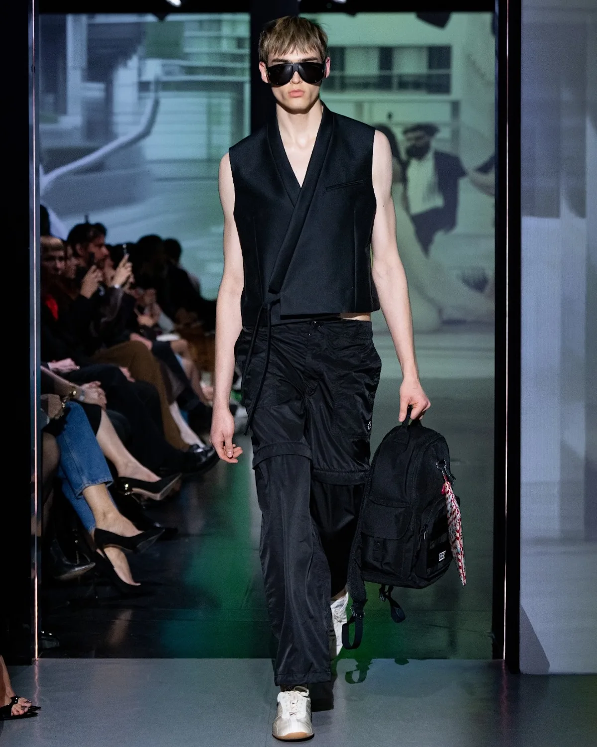 Цей виріб демонструє сучасний мінімалістичний стиль у чорному кольорі – безрукавний топ у стилі блейзера поєднується з вільними штанами та кросівками. Великі сонцезахисні окуляри та рюкзак доповнюють образ, надаючи йому урбаністичного, функціонального вигляду.