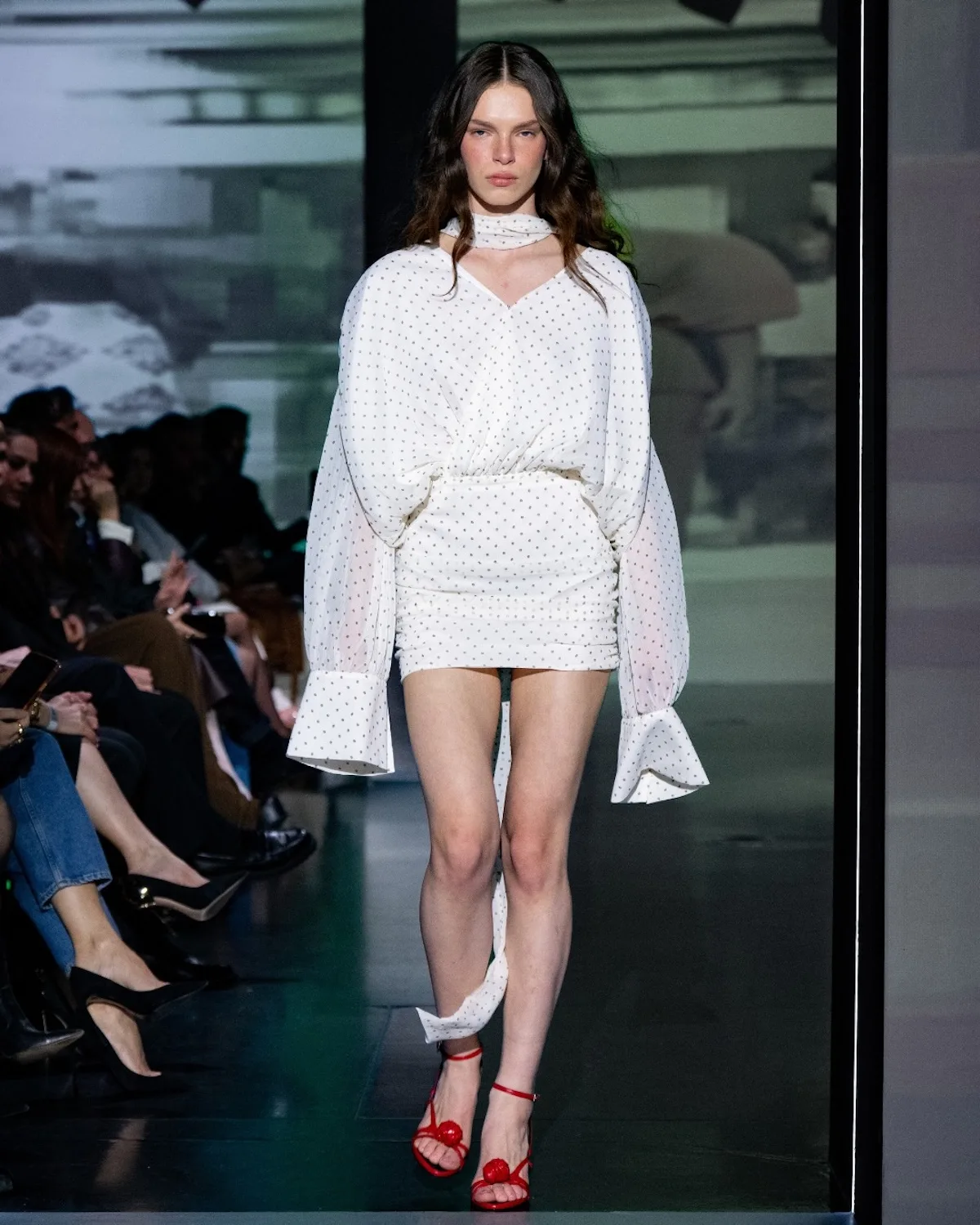 Модель пройшлася подіумом у легкій білій міні-сукні в ніжний горошок, з пишними рукавами та тонкою зав'язкою на шиї. Образ доповнюють червоні босоніжки на високих підборах, які додають контрасту та підкреслюють жіночний, свіжий характер вбрання.