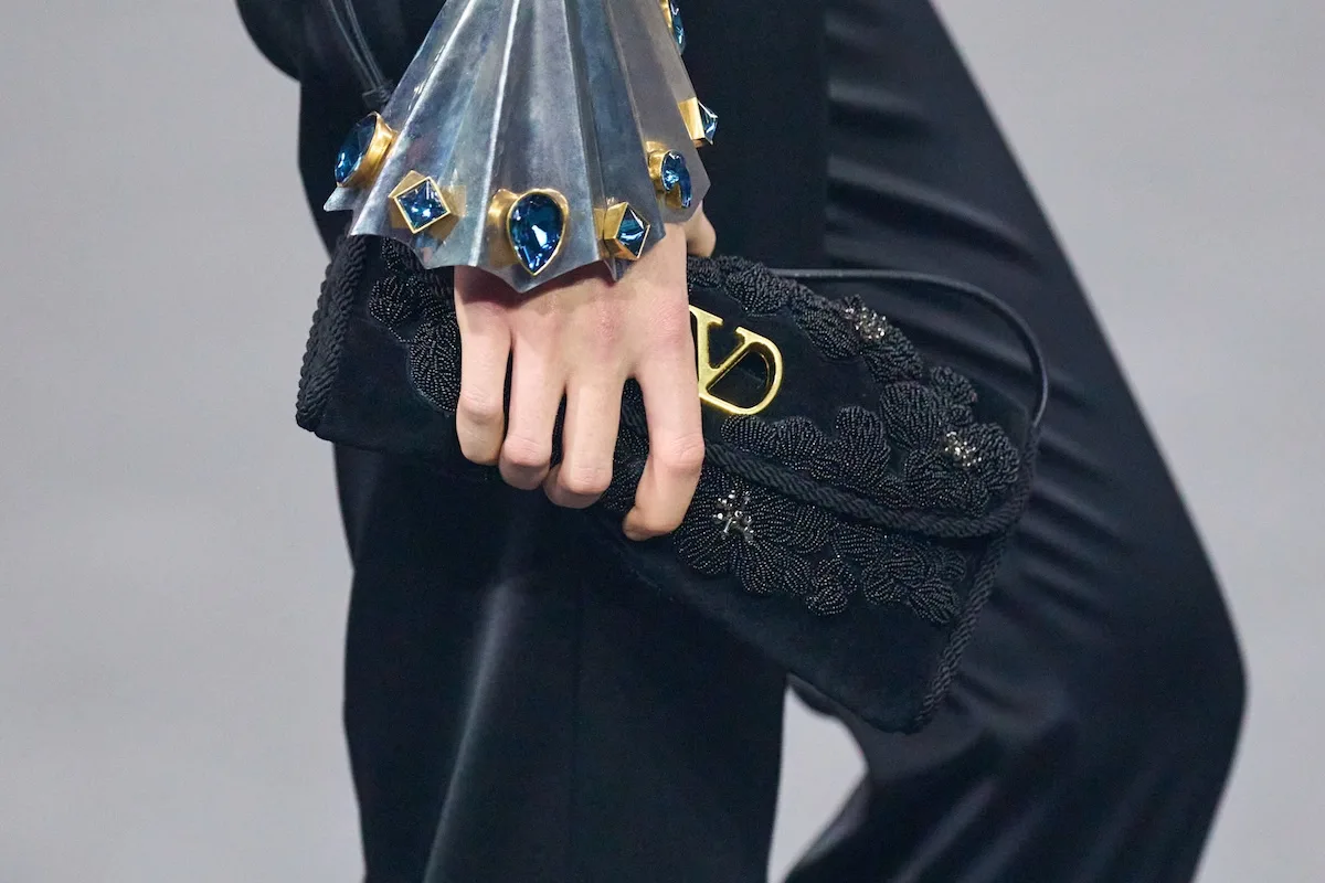 Крупний план чорного, прикрашеного клатча Valentino, який тримає в руці модель, з помітним золотим логотипом та вишукано оздобленою манжетою з синім камінням. Елегантна деталь підкреслює розкішний характер бренду, який також зіткнувся з юридичною перевіркою за порушення в ланцюжку поставок. Контраст між естетикою продукту та закулісним виробництвом служить нагадуванням про важливість прозорості в преміальній індустрії.