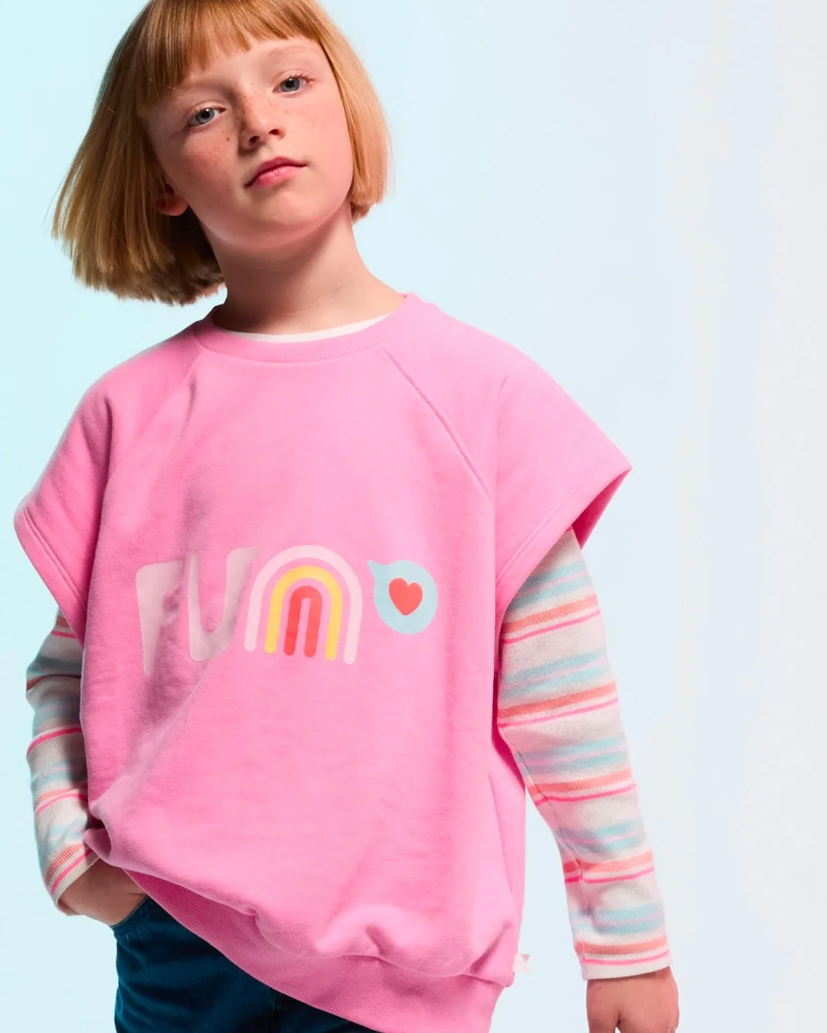 Дівчина в рожевій толстовці з яскравим принтом та довгими смугастими рукавами представляє повсякденний дитячий одяг. Вбрання легке, яскраве та підходить для щоденного носіння. Фото відображає характер дитячої модної колекції Okaïdi.